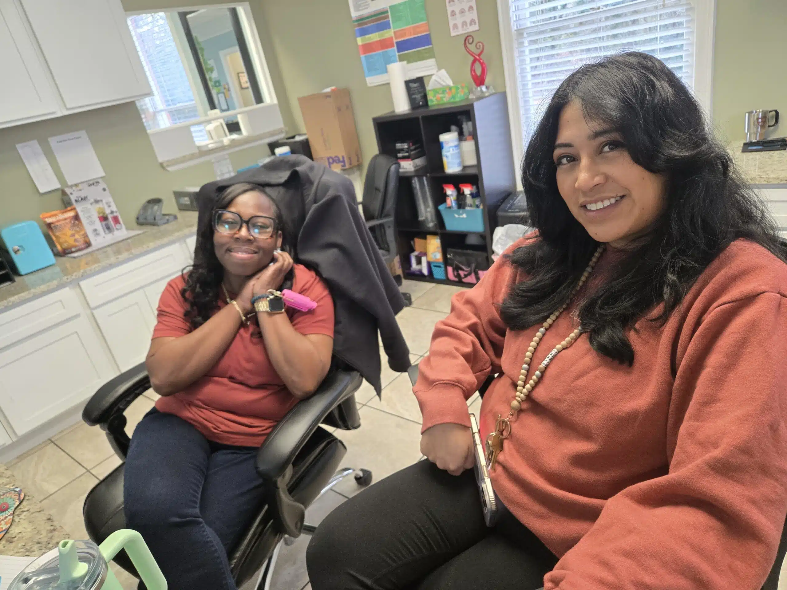 Two women sit smiling in an office, both wearing reddish tops. One is sitting back with her hands by her face, while the other leans forward holding a phone. Office supplies and shelves are visible in the background.