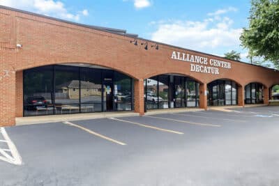 A brick building with arched windows labeled Alliance Center Decatur, facing an empty parking lot under a partly cloudy sky.