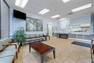 A clean, empty waiting room with rows of chairs, a coffee table, a large plant, artwork on the wall, tiled floor, a TV mounted high, and a reception window on the right.