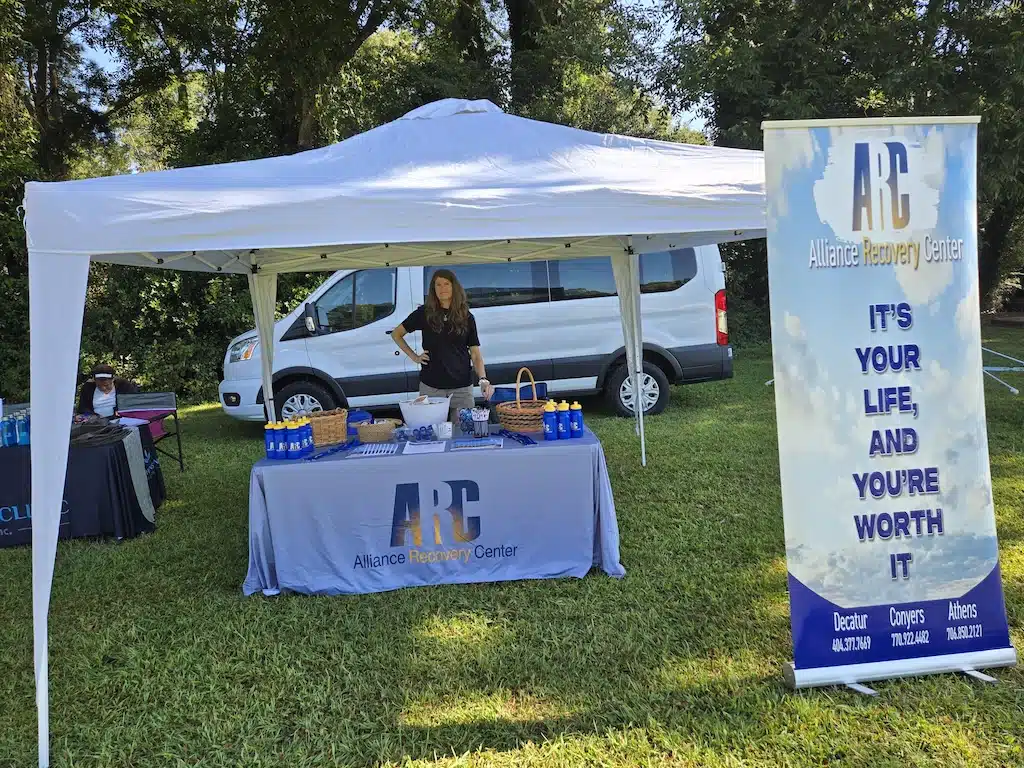 A woman stands at an Alliance Recovery Center booth under a white tent, with informational items on the table and a large banner that reads, Its your life, and youre worth it. A white van is parked behind the tent.