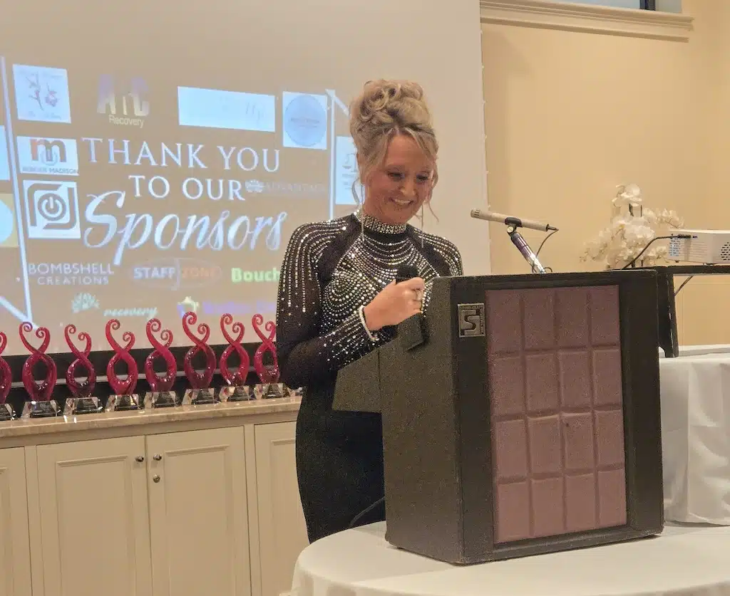 A woman in a sparkly black dress speaks at a podium during an event, with a screen behind her displaying Thank you to our sponsors and various sponsor logos.