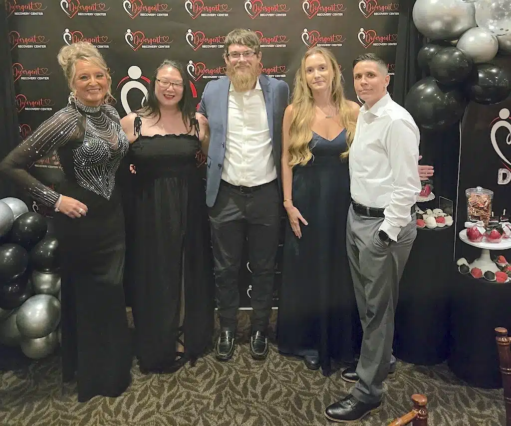 Five people dressed formally stand together at an event with a black and red backdrop, balloons, and a dessert table. They are smiling and posing for the photo on a patterned carpet.