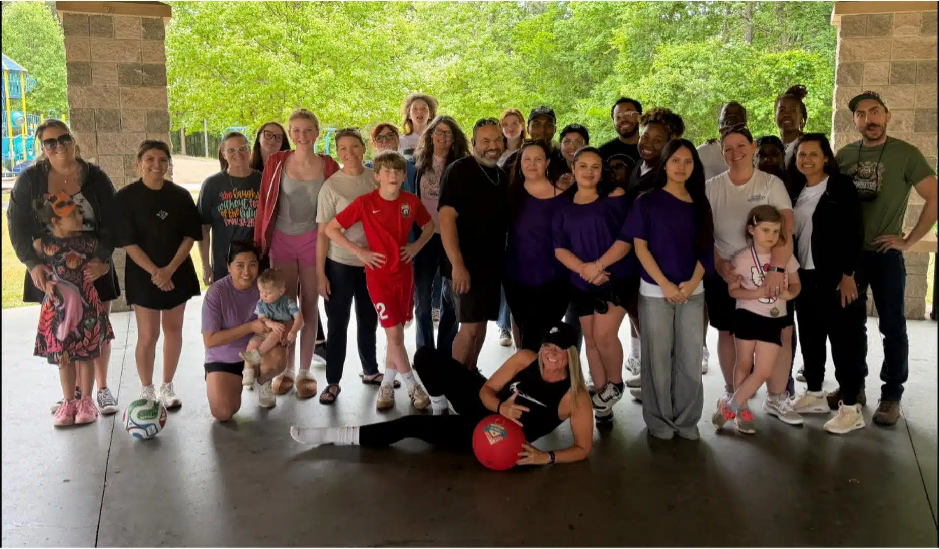 Field Day A diverse group of adults and children pose together under a pavilion, some wearing purple and sportswear. Trees and a playground are visible in the background. One person in front is leaning on a large red ball.
