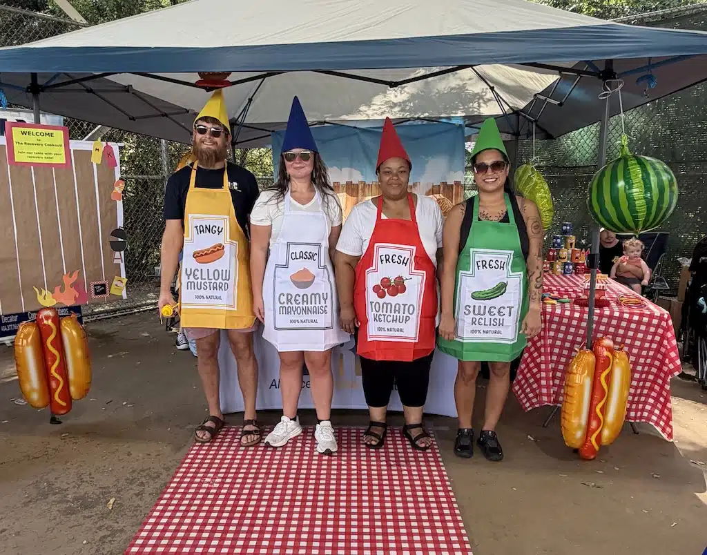 Four adults stand under a canopy, each wearing an apron and hat styled as mustard, mayonnaise, ketchup, and relish bottles. Picnic tables and hot dog decorations are visible around them.