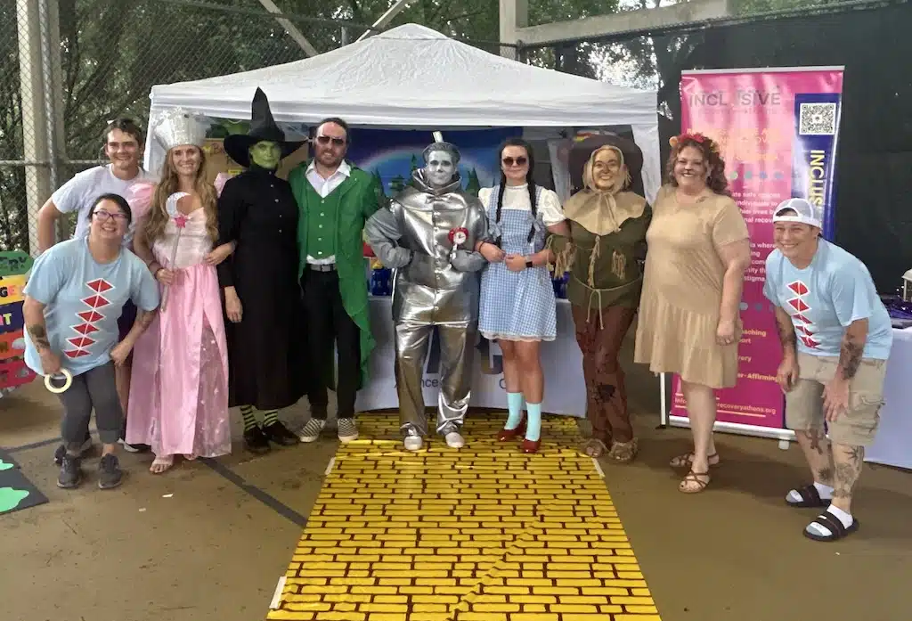 A group of people in Wizard of Oz costumes stand on a yellow brick road in front of a tent at an outdoor event, smiling at the camera.