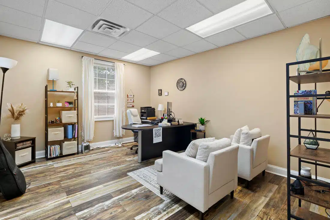 Therapy2 Modern office with beige walls, wood floor, a desk with computer, two white armchairs, shelves with decor, a large window with blinds, and ceiling lights.