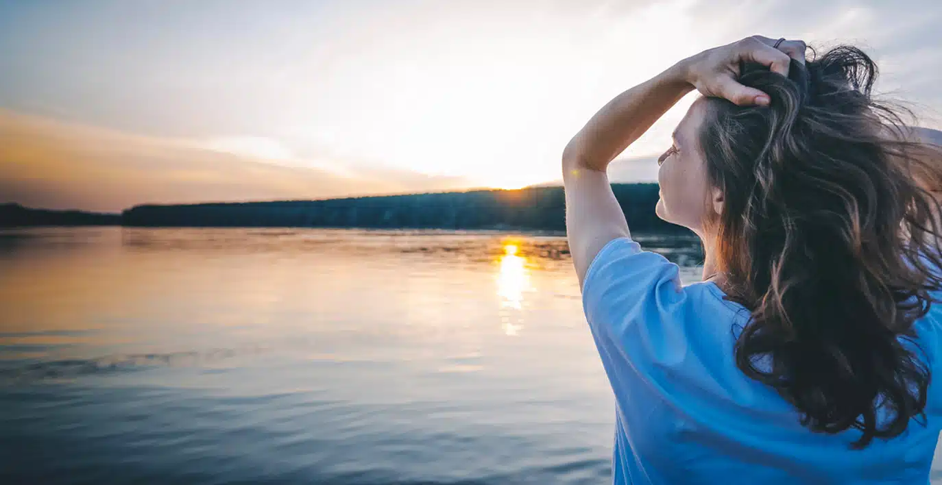 A person with long hair stands by a calm lake at sunset, holding their hair back and gazing at the water and distant treeline.