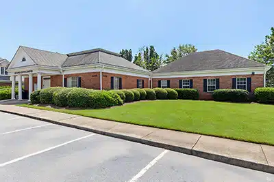 A single-story brick building with white trim, surrounded by neatly trimmed bushes and a well-maintained lawn, with a parking lot in the foreground under a clear, sunny sky.