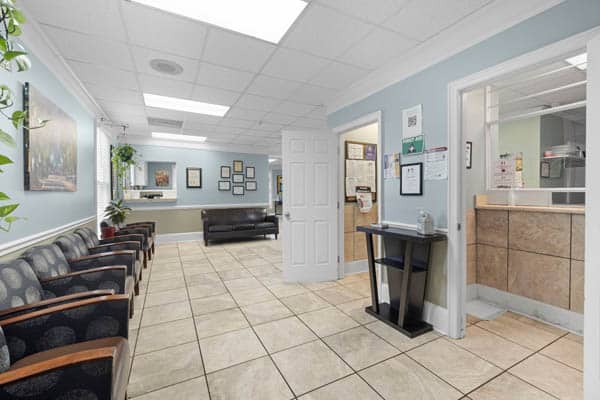 athens-arc-facility-3 A clean, bright waiting room with patterned chairs along one wall, a black couch, tiled floor, wall art, and two open doors leading to office areas. A small table with sanitizer is near the doorway.
