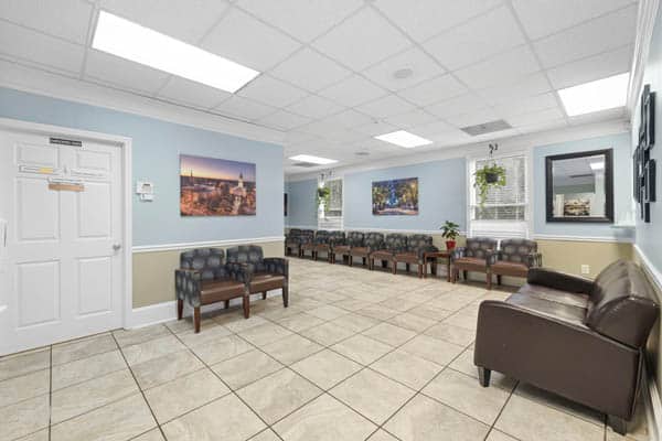 athens-arc-facility-4 Spacious, well-lit waiting room with rows of chairs, a brown leather couch, light blue walls, tile flooring, and nature photos on the walls. Large windows let in natural light.