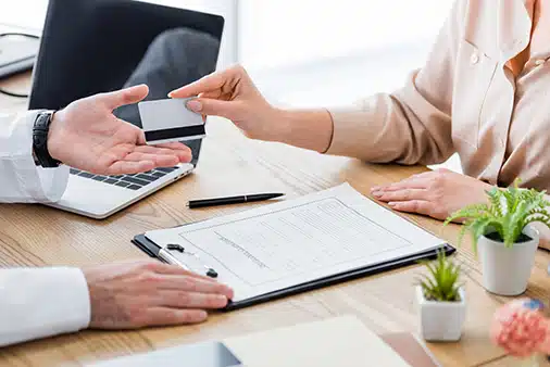 Two people sit at a desk. One person hands a credit card to the other. A clipboard with a form, a laptop, pen, and small potted plants are on the table.