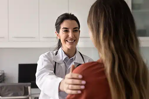 A smiling doctor in a white coat gently rests a hand on a patients shoulder, offering comfort and reassurance in a medical office setting.