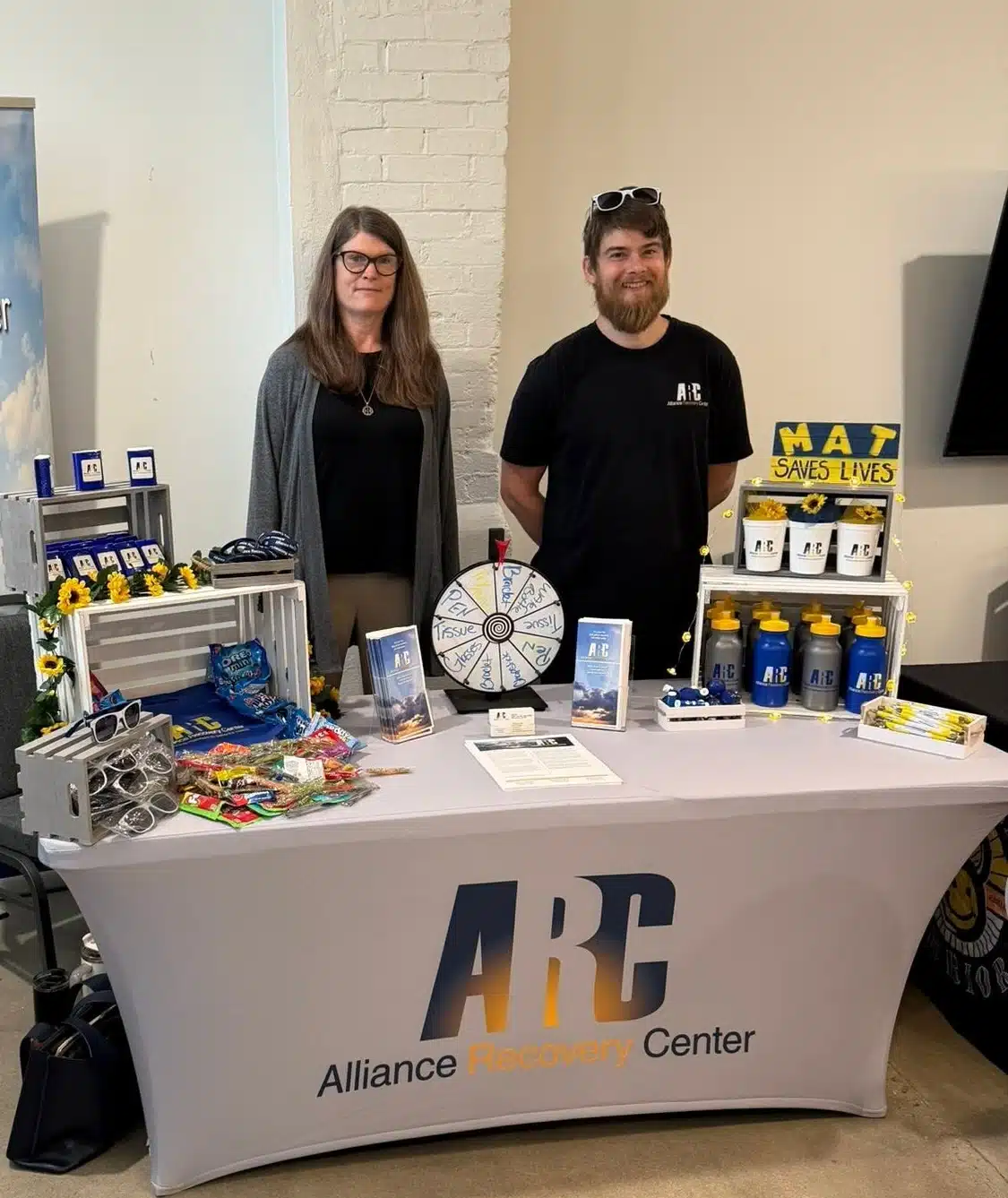 Two people stand behind an Alliance Recovery Center table with brochures, prizes, branded items, and a prize wheel. The display promotes MAT and substance recovery resources. Both are smiling at the camera.